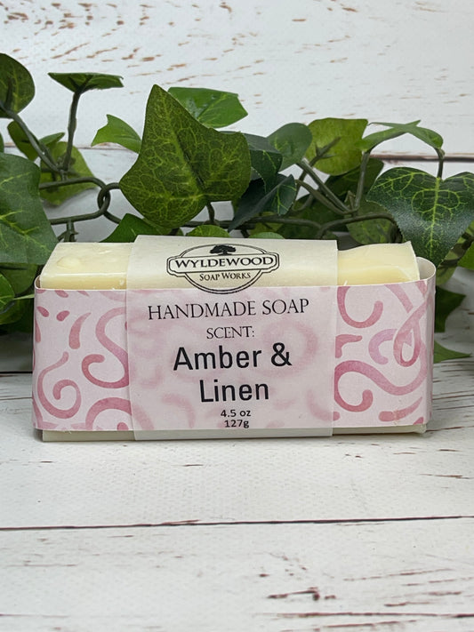 A light cream colored bar of scented goat milk soap named Amber and Linen wrapped with pink scrapbook paper and a vellum label on a white back ground with green leaves behind in the background