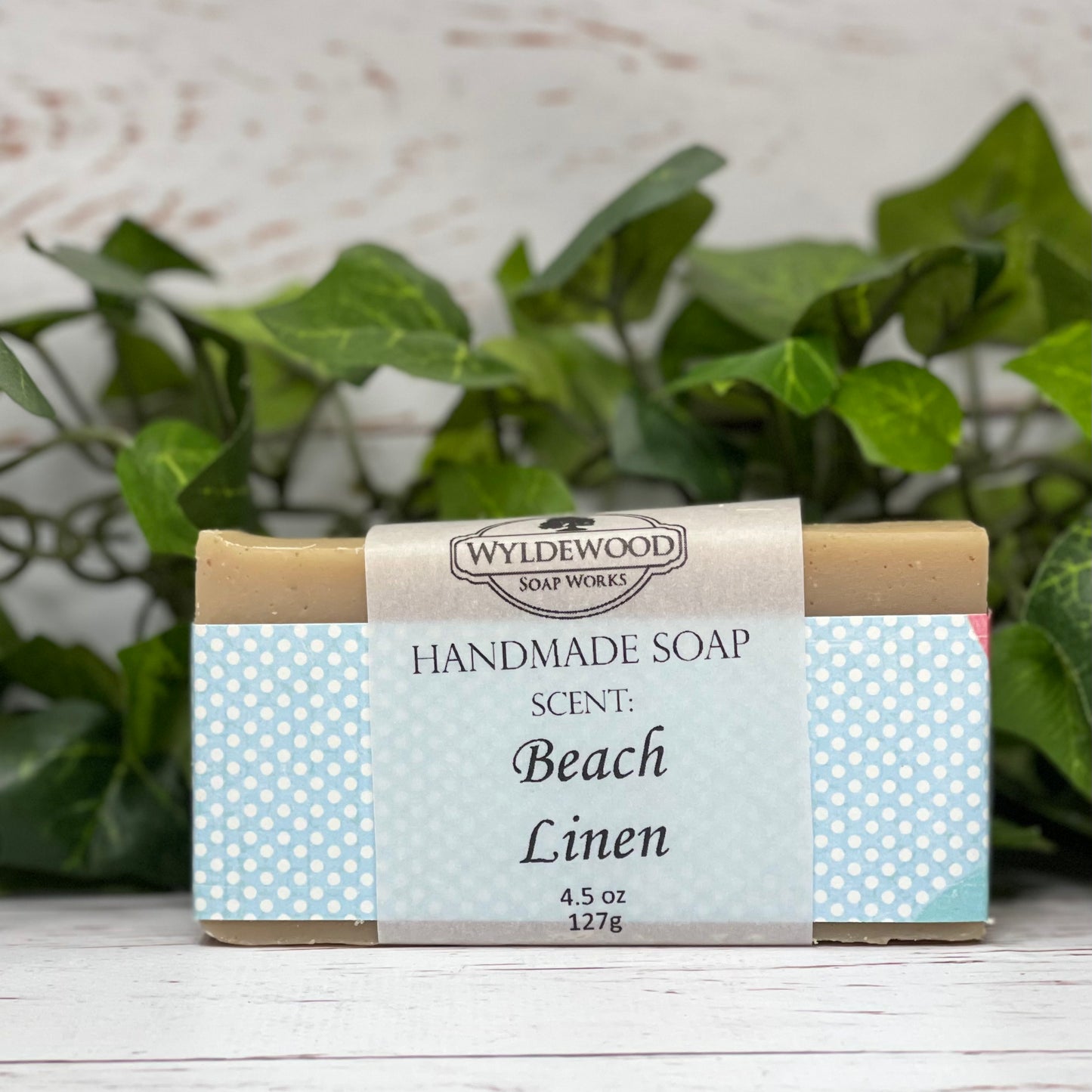A bar of handmade soap with packaging that has 'WYLDEWOOD HANDMADE SOAP' and the scent listed as 'Beach Linen'. The bar is placed on a white surface with leaves in the background.