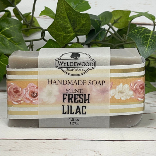 A light purple gray bar of Fresh Lilac scented goat milk soap wrapped with floral scrapbook paper and a vellum label on a white back ground with green leaves behind