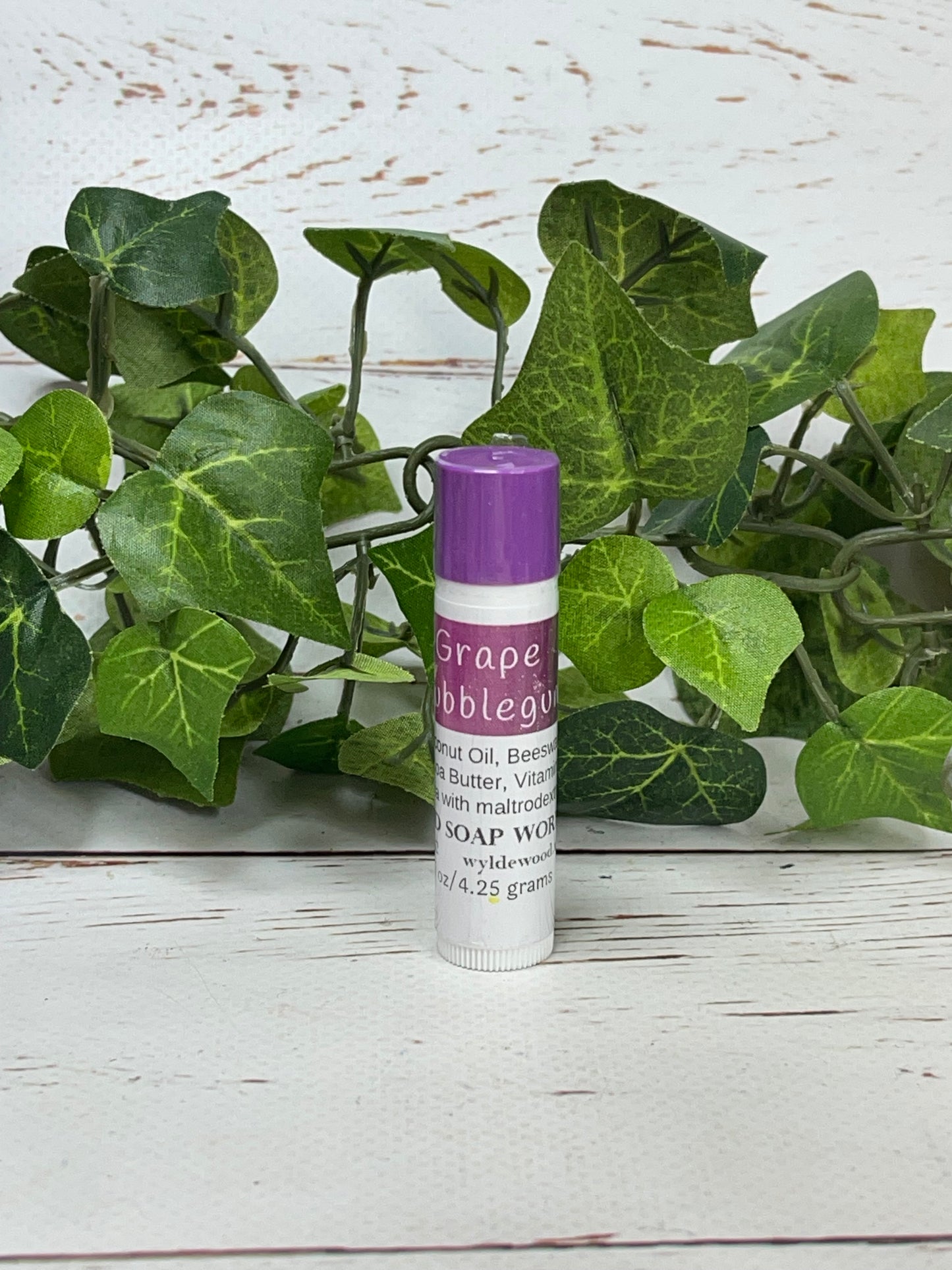 A tube of lip balm standing on a white background labelled Grape Bubblegum flavor with green leaves behind the tube of lip balm