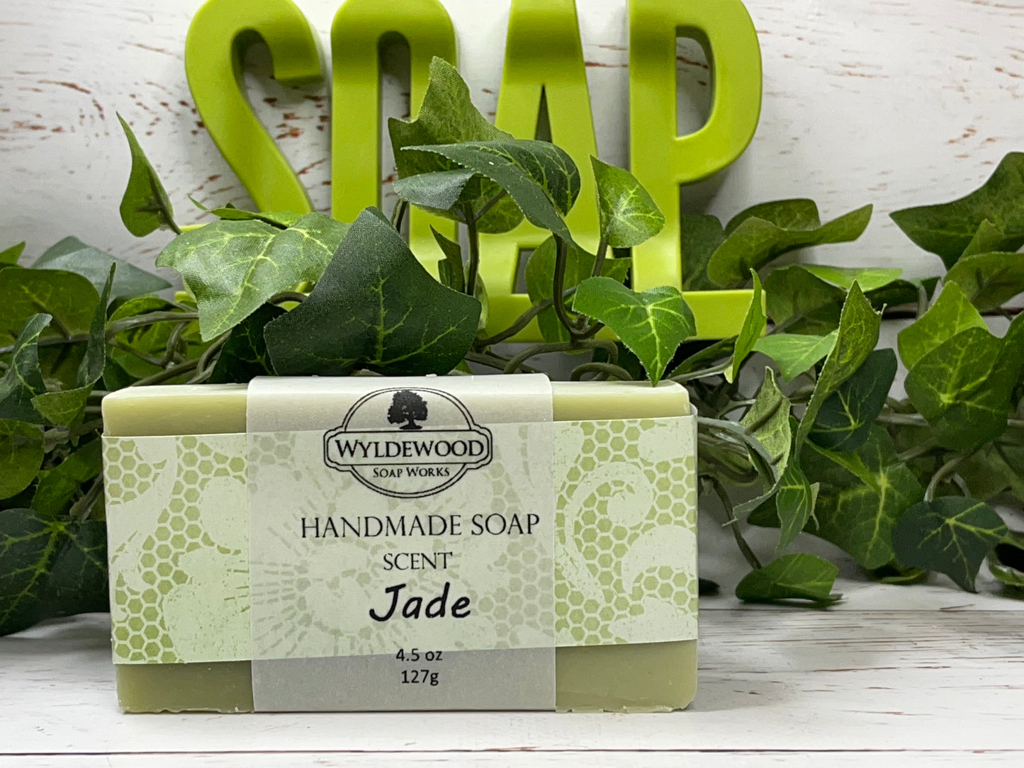 A bar of handmade soap with packaging that has 'WYLDEWOOD HANDMADE SOAP' and the scent listed as 'Jade'. The bar is placed on a white surface with leaves in the background.