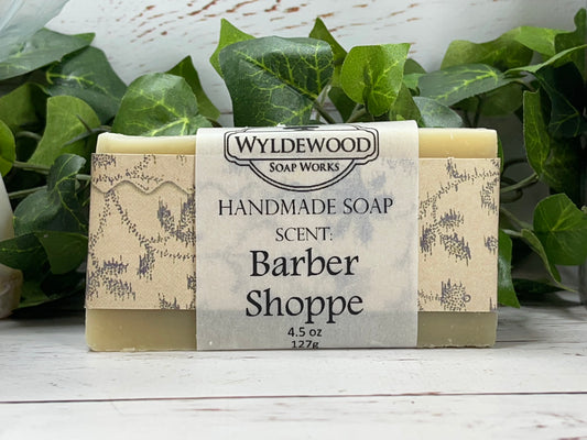 A bar of handmade soap with packaging that has 'WYLDEWOOD HANDMADE SOAP' and the scent listed as 'Barber Shoppe'. The bar is placed on a white surface with leaves in the background.