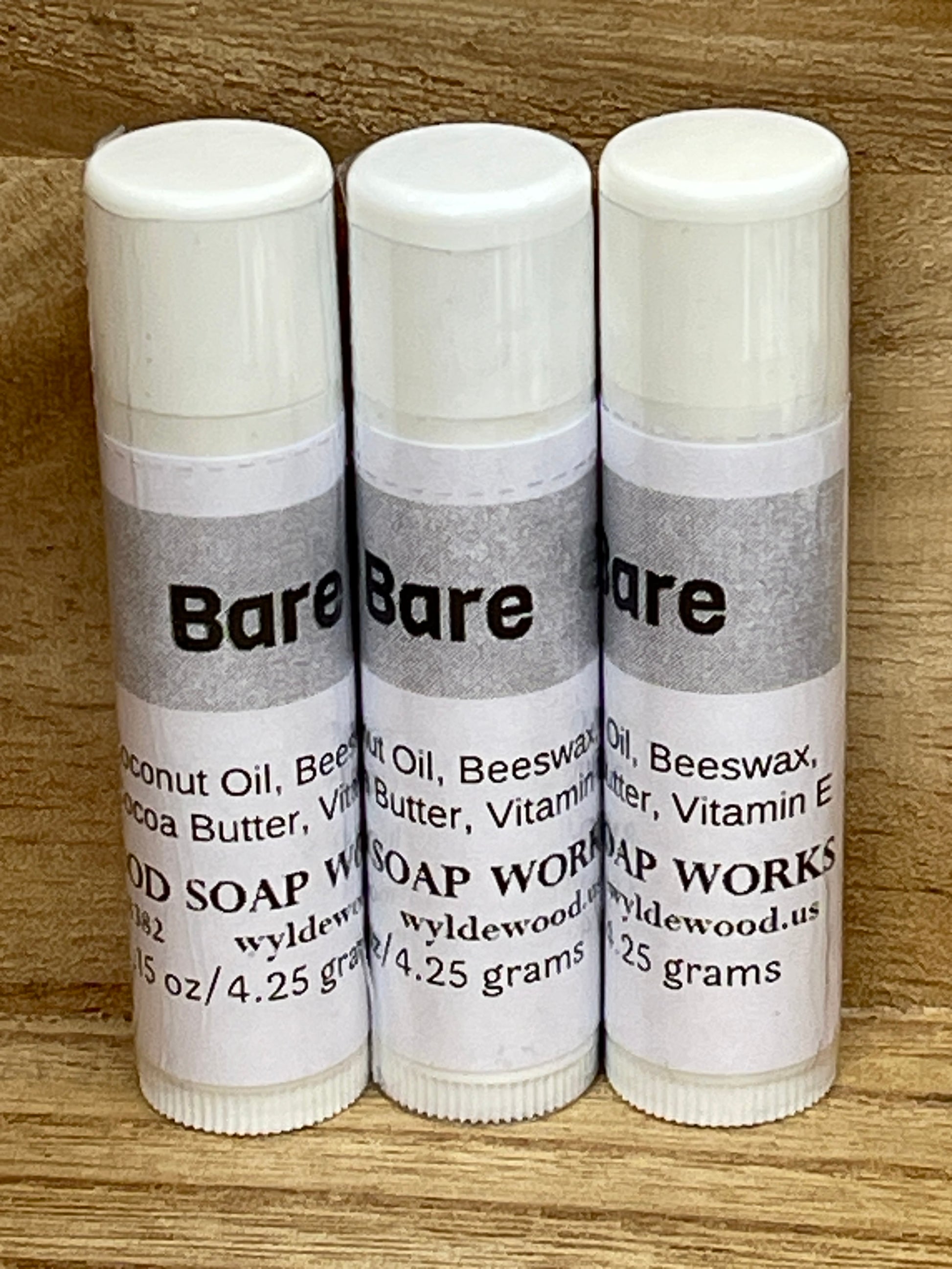 Three tubes of Bare unflavored lip balms made by Wyldewood soap works standing on a wood background.