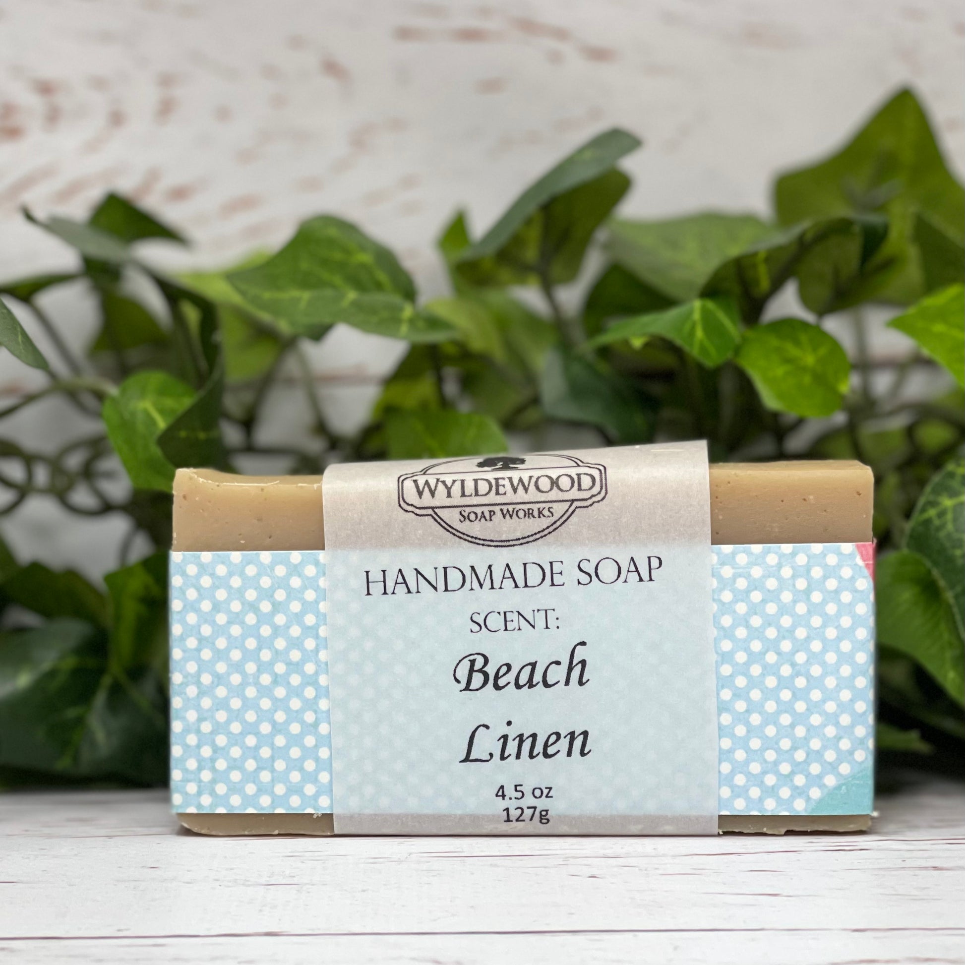 A bar of handmade soap with packaging that has 'WYLDEWOOD HANDMADE SOAP' and the scent listed as 'Beach Linen'. The bar is placed on a white surface with leaves in the background.