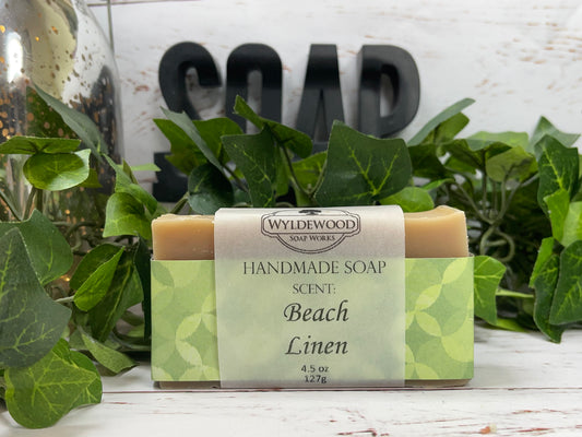 A bar of handmade goat milk soap with 'BEACH LINEN' scent, packaged in a clear wrapper with a green and white label, displayed on a white surface with greenery in the background.