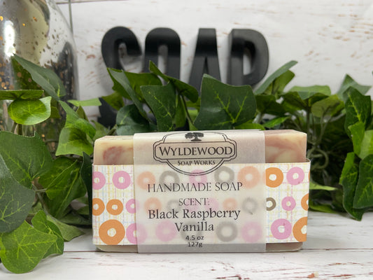 A bar of handmade goat milk soap with 'Saturday Morning' text and cereal illustration on the packaging, placed on a wooden surface with green leaves and a 'SOAP' sign in the background.