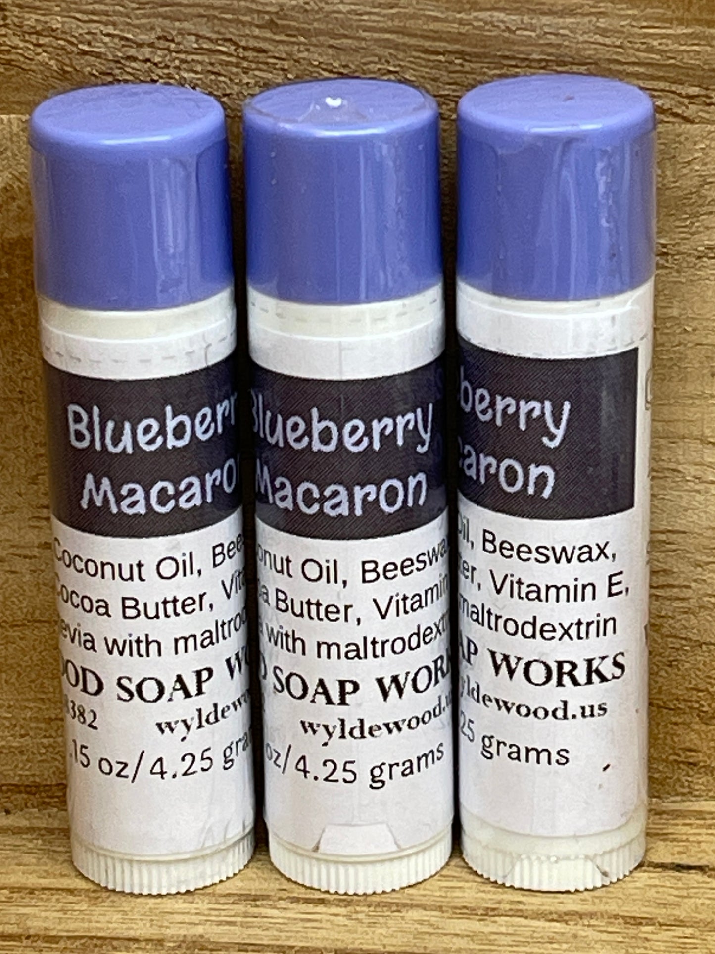 Three tubes of Blueberry Macaron flavor lip balms made by Wyldewood soap works standing on a wood background.