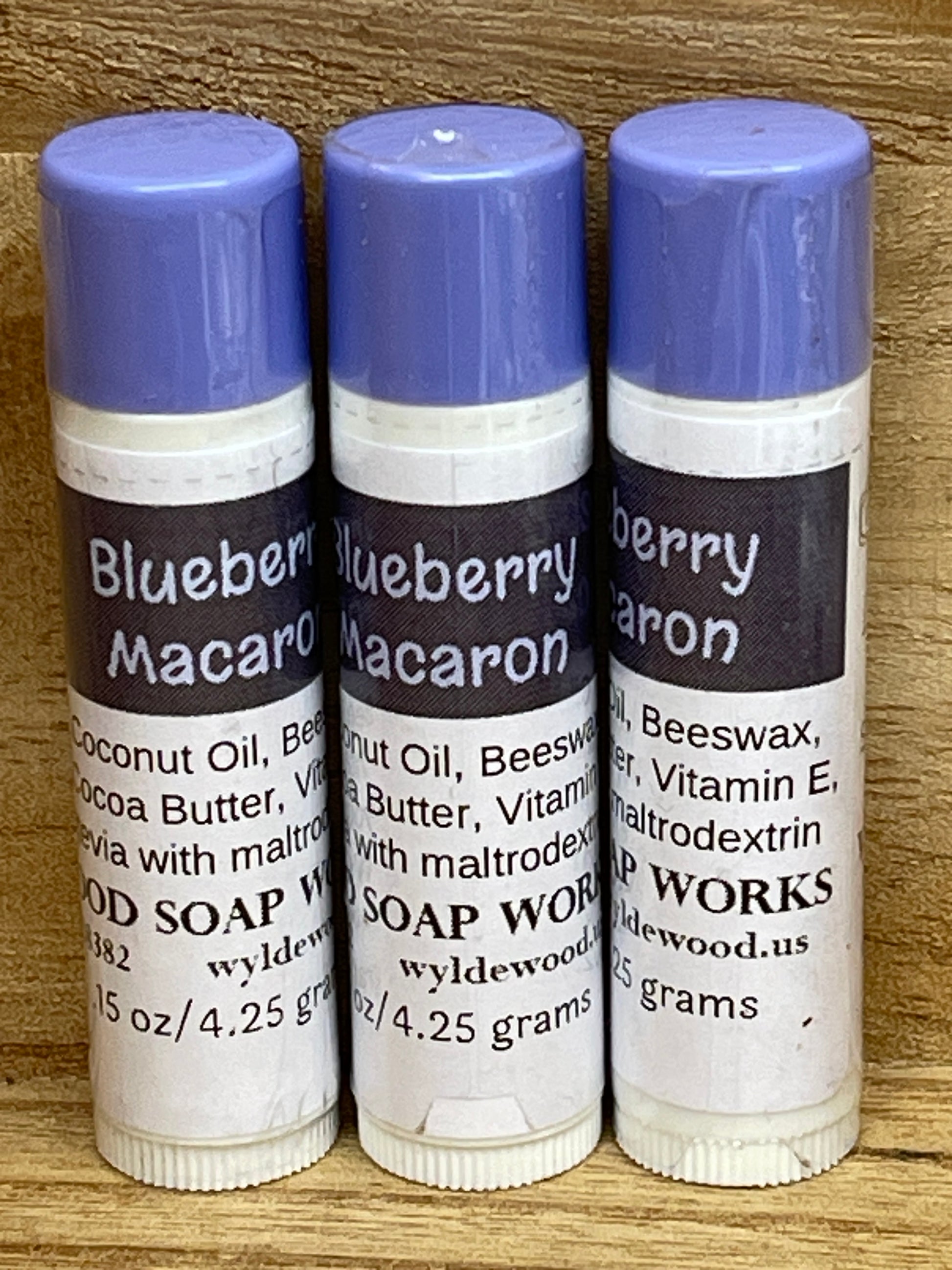 Three tubes of Blueberry Macaron flavor lip balms made by Wyldewood soap works standing on a wood background.