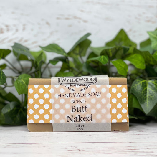 A bar of handmade soap with packaging that has 'WYLDEWOOD HANDMADE SOAP' and the scent listed as 'Butt Naked'. The bar is placed on a white surface with leaves in the background.