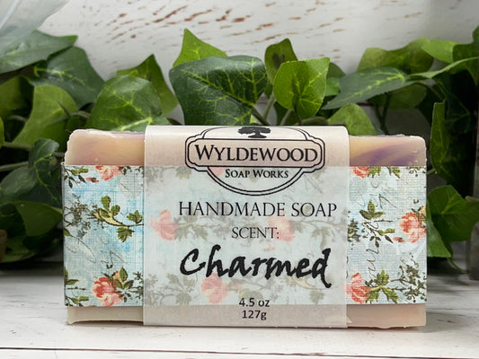 A bar of handmade scented soap with goat milk, packaged in a floral-designed wrapper with 'Wyldewood Soap Works' branding and the word 'Charmed' on it, set against a natural background.