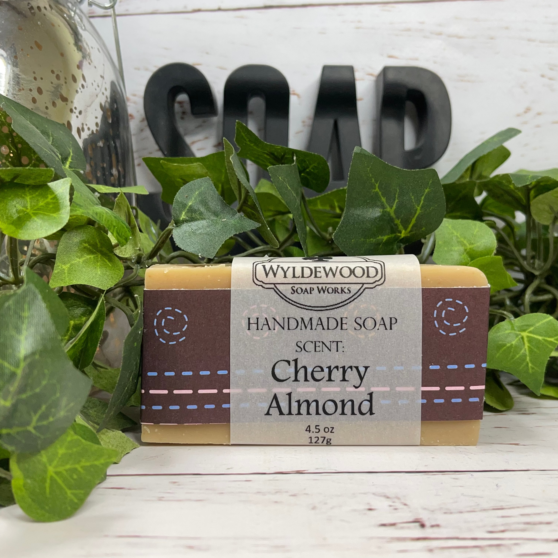 A bar of handmade soap with 'Wyldewood Handmade Soap, Cherry Almond' printed on the label, resting on a white wooden surface with green leaves around it.