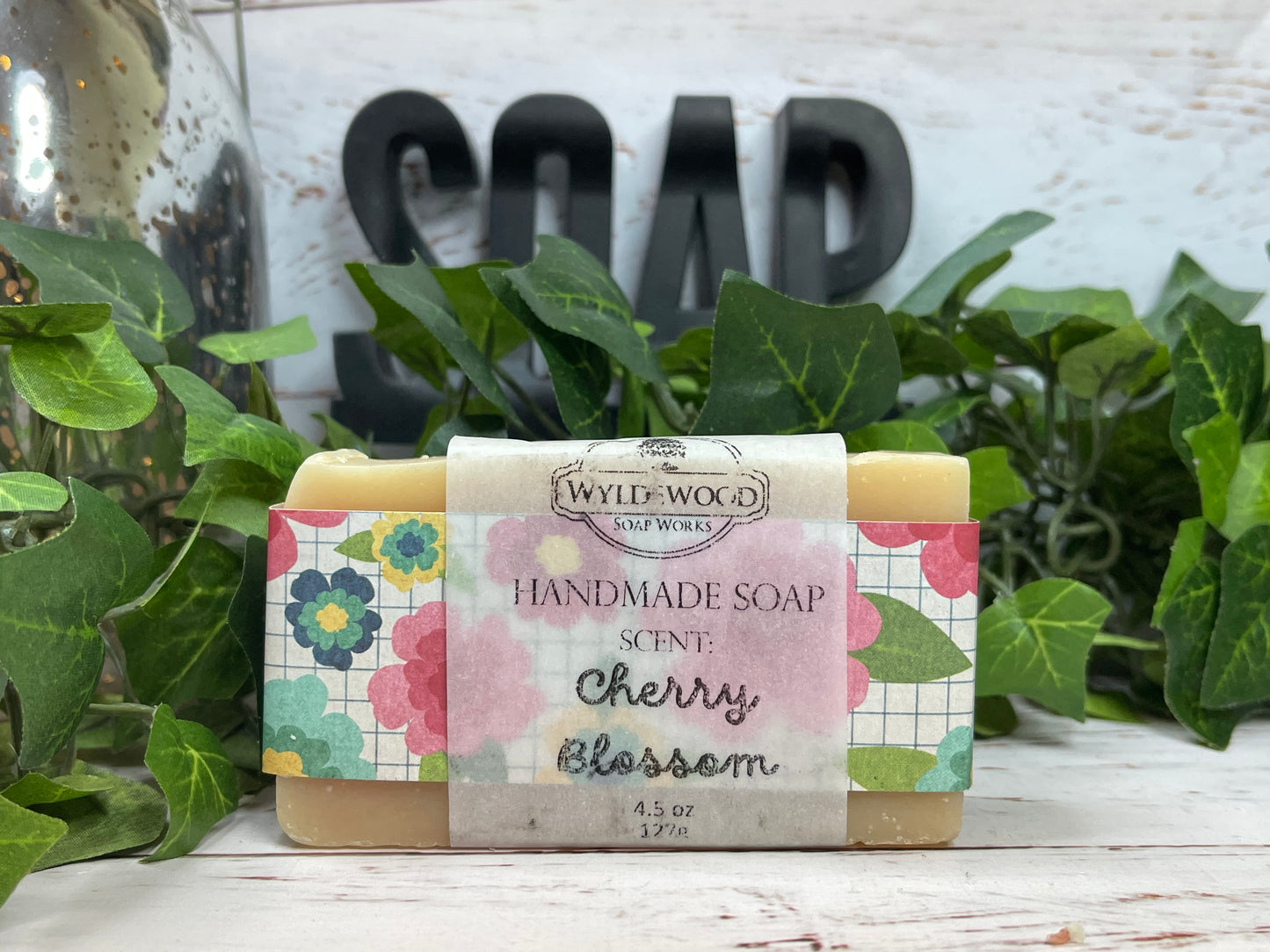 A bar of handmade goat milk soap with 'Cherry Blossom' labeled on it, surrounded by green leaves and a 'SOAP' sign in the background.