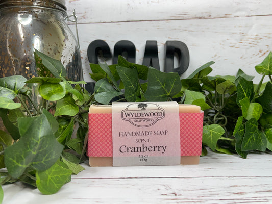 A bar of handmade goat milk soap with 'CRANBERRY' printed on the label, surrounded by green leaves and a jar in the background, on a wooden surface.