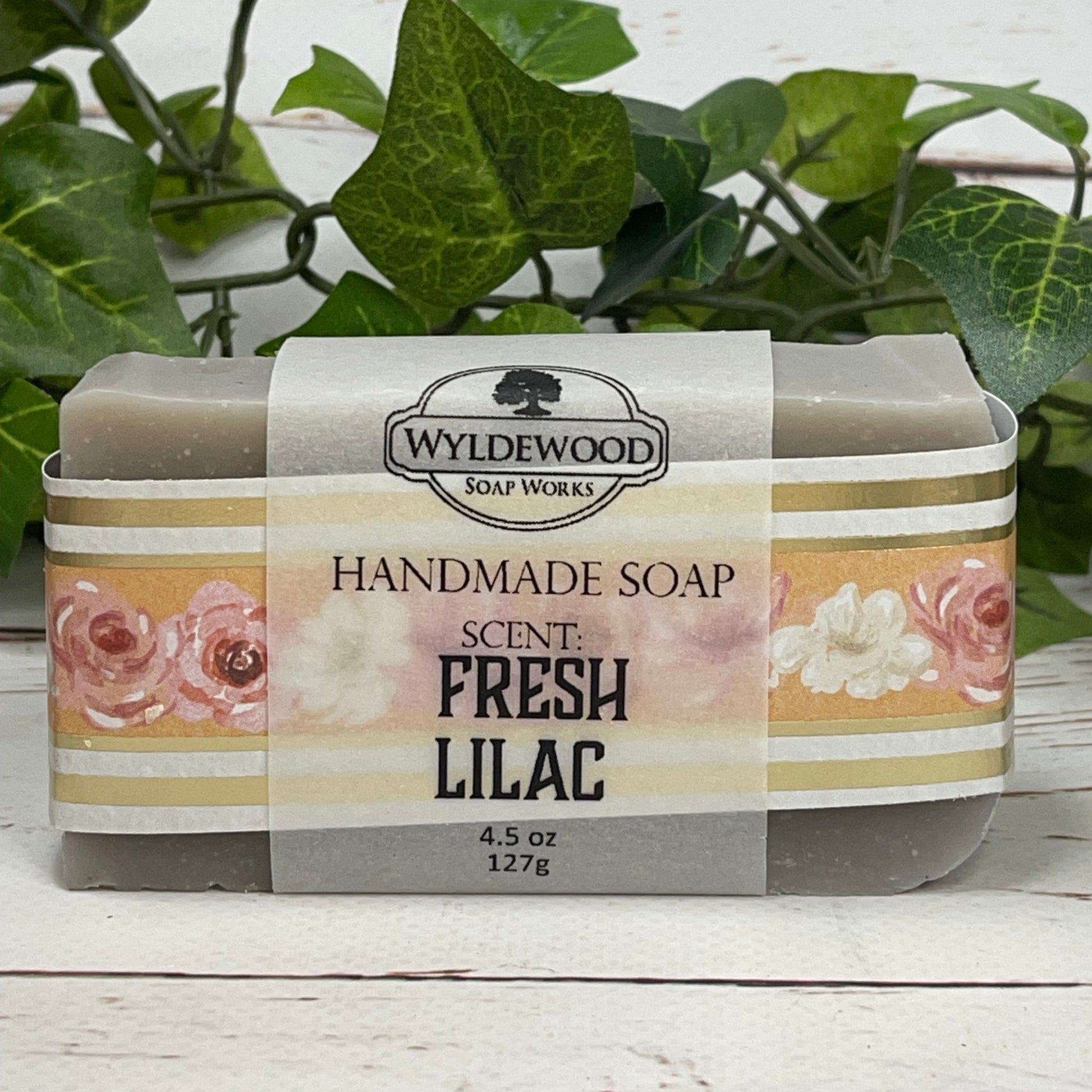 A light purple gray bar of Fresh Lilac scented goat milk soap wrapped with floral scrapbook paper and a vellum label on a white back ground with green leaves behind