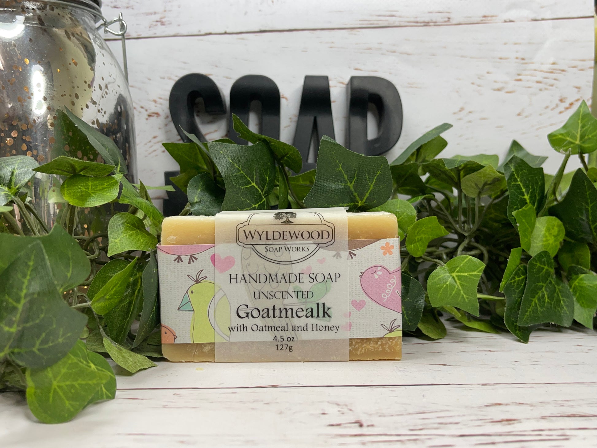 Bar of handmade goat milk soap with 'Wyldewood' branding and 'Goatmealk with Oatmeal and Honey' text, placed on a wooden surface surrounded by green leaves and a jar in the background