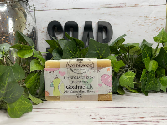 Bar of handmade goat milk soap with 'Wyldewood' branding and 'Goatmealk with Oatmeal and Honey' text, placed on a wooden surface surrounded by green leaves and a jar in the background