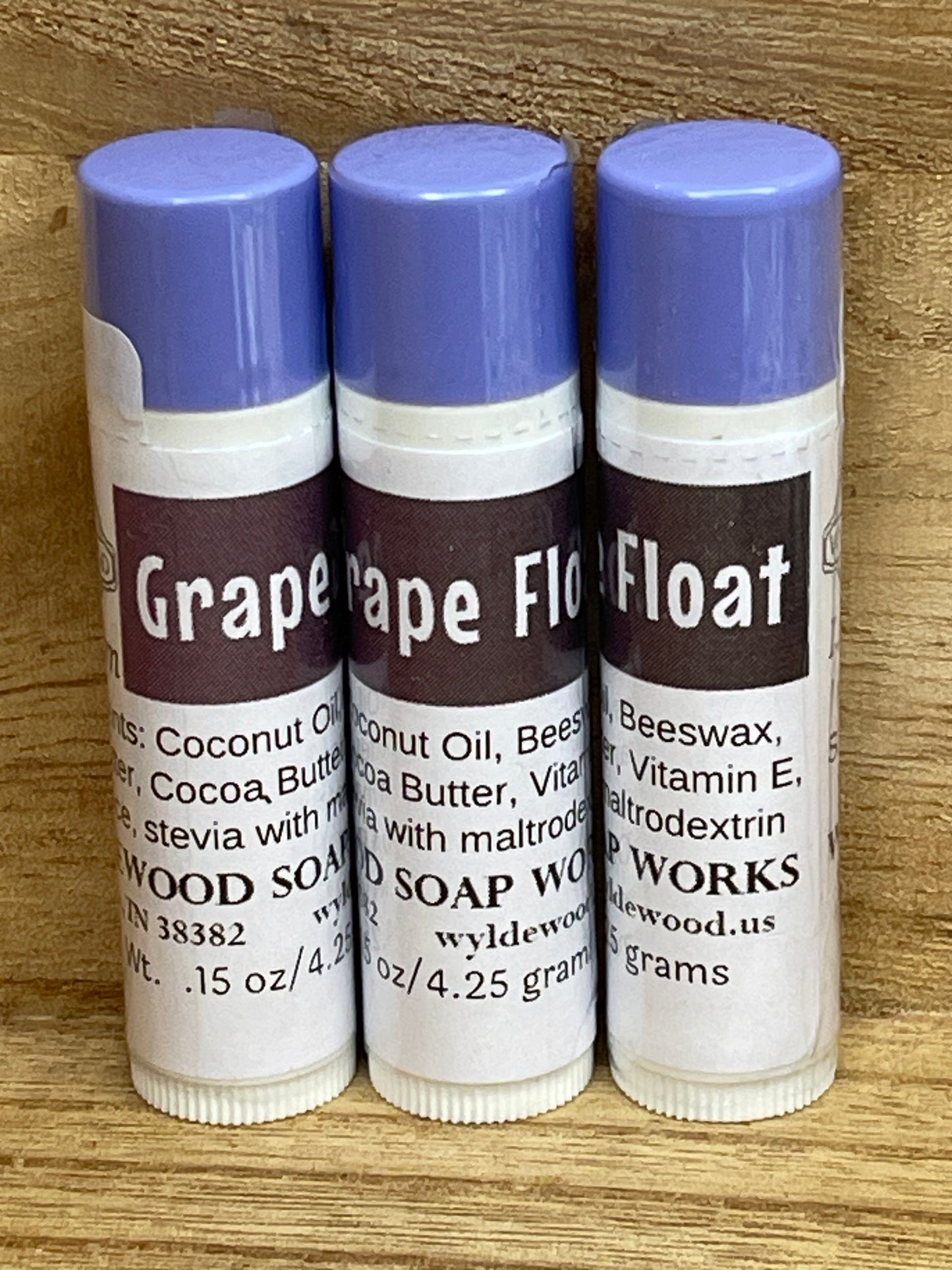Three tubes of Grape Float flavor lip balms made by Wyldewood soap works standing on a wood background.