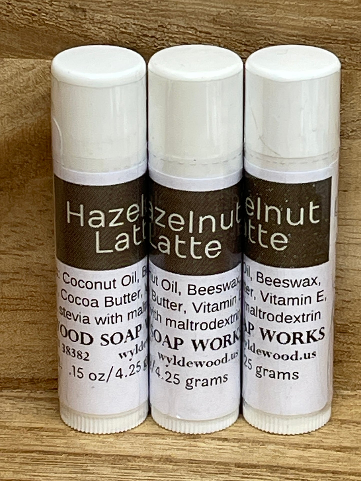 Three tubes of Hazelnut Latte flavor lip balms made by Wyldewood soap works standing on a wood background.