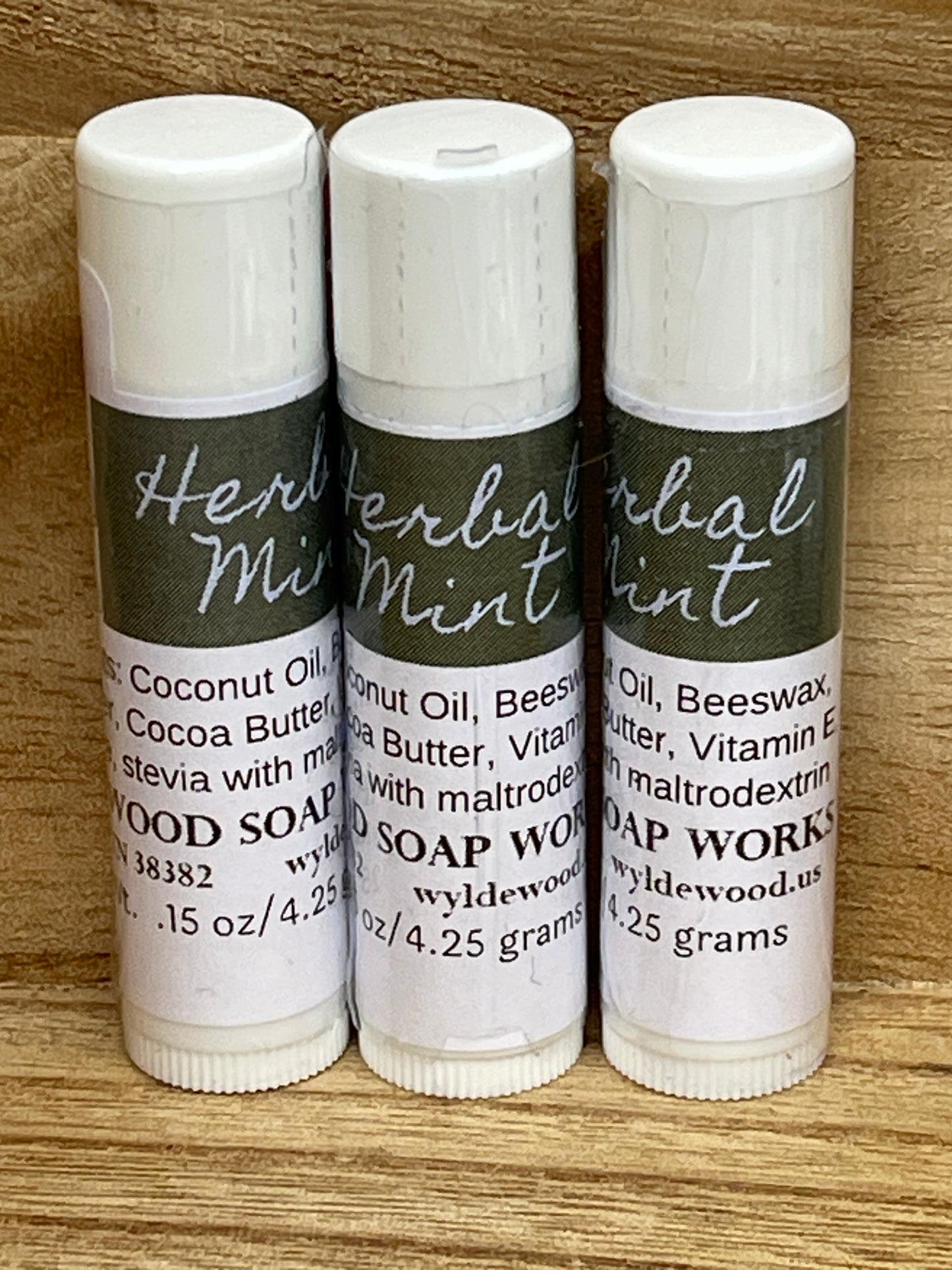 Three tubes of Herbal Mint flavor lip balms made by Wyldewood soap works standing on a wood background.