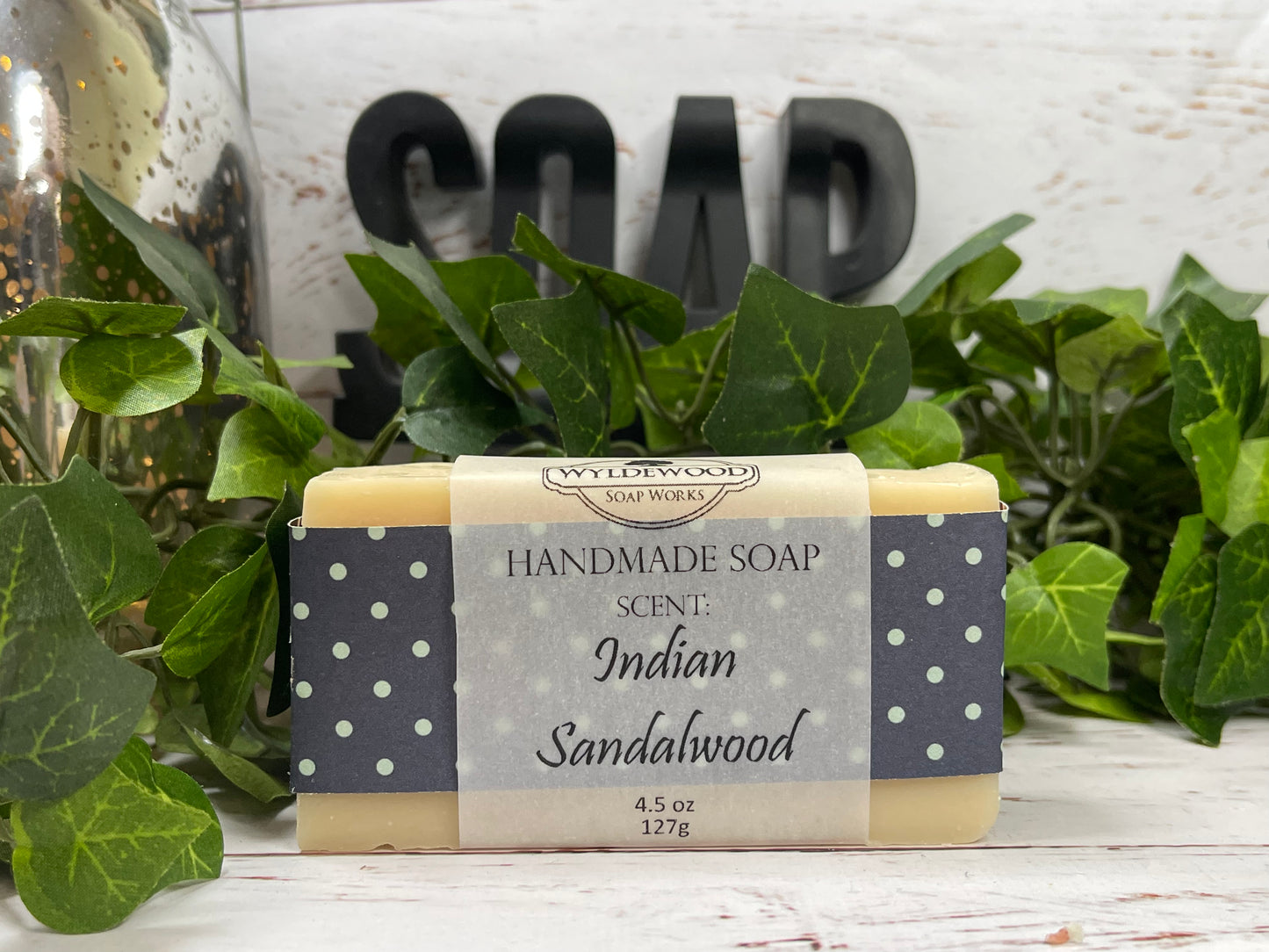 A bar of handmade goat milk soap with 'Indian Sandalwood' scent, placed on a white surface with green leaves and a 'SOAP' sign in the background.