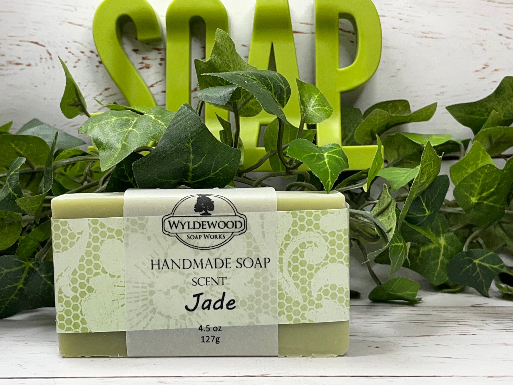 A bar of handmade soap with packaging that has 'WYLDEWOOD HANDMADE SOAP' and the scent listed as 'Jade'. The bar is placed on a white surface with leaves in the background.
