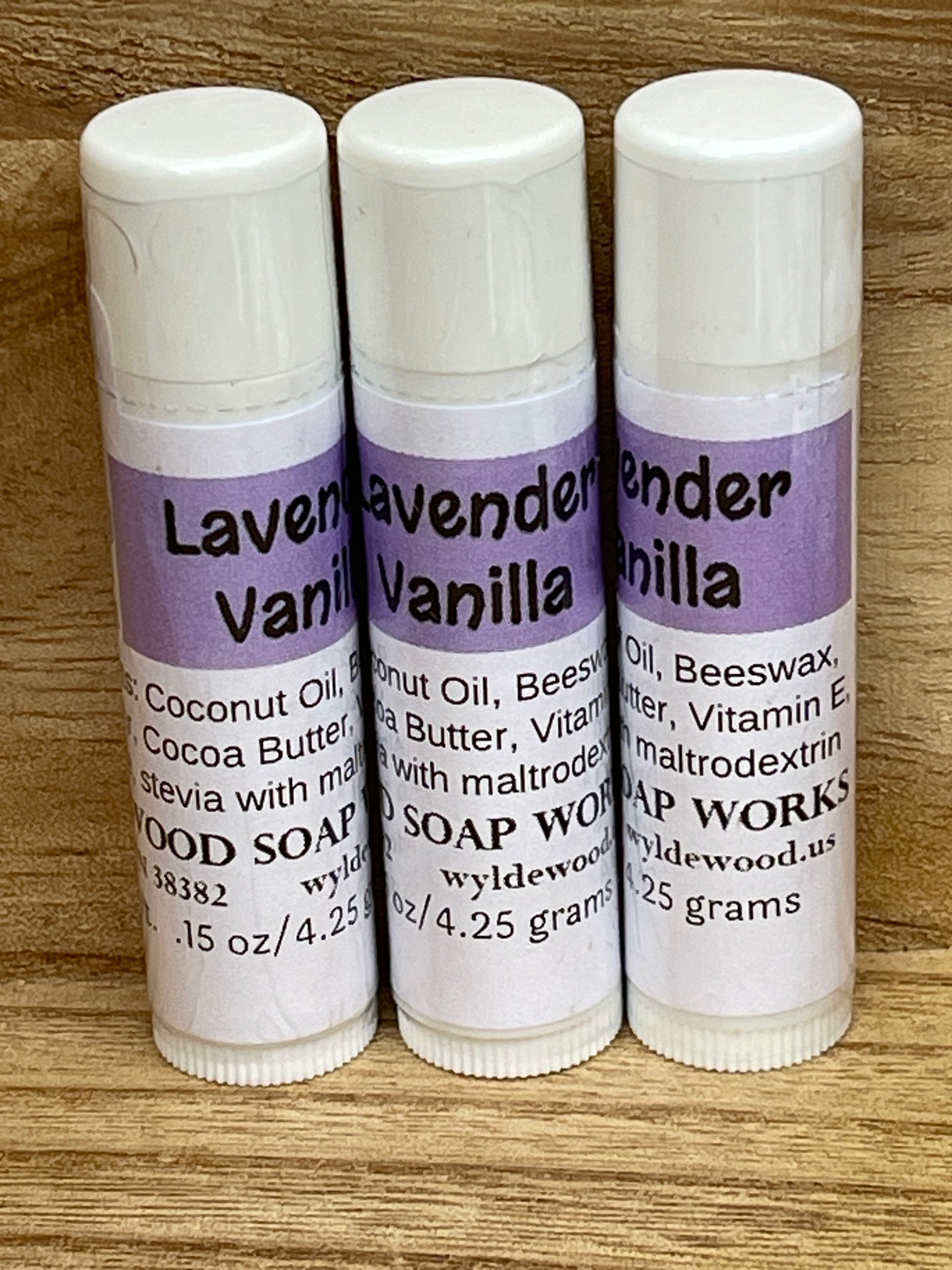 Three tubes of Lavender Vanilla flavor lip balms made by Wyldewood soap works standing on a wood background.