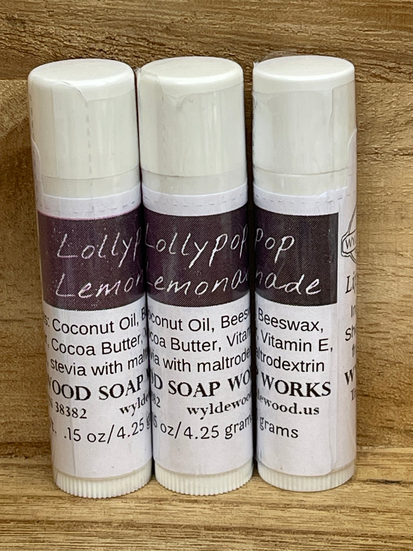 Three tubes of Lollypop Lemonade flavor lip balms made by Wyldewood soap works standing on a wood background.