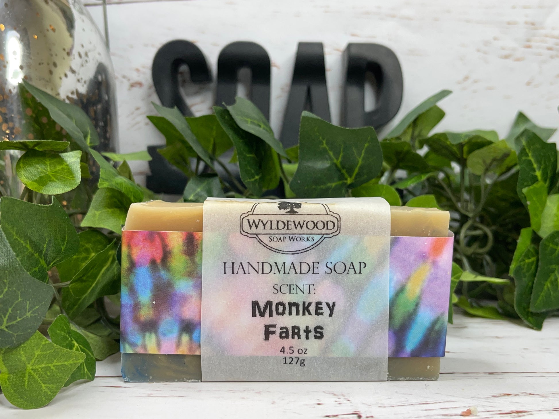 A bar of handmade goat milk soap with 'Monkey Farts' labeled on the packaging, placed on a white surface with green leaves and a 'SOAP' sign in the background.