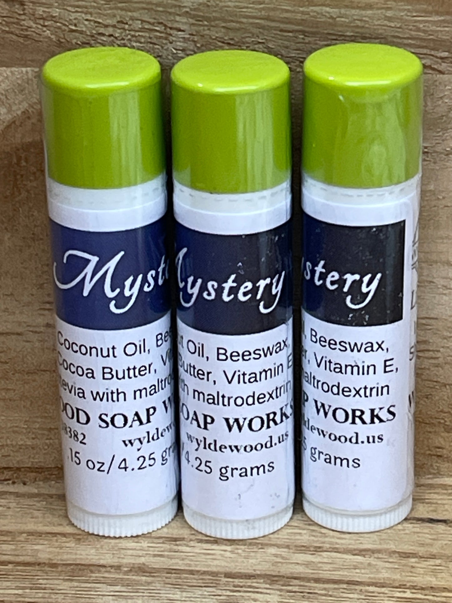 Three tubes of Mystery flavor lip balms made by Wyldewood soap works standing on a wood background.