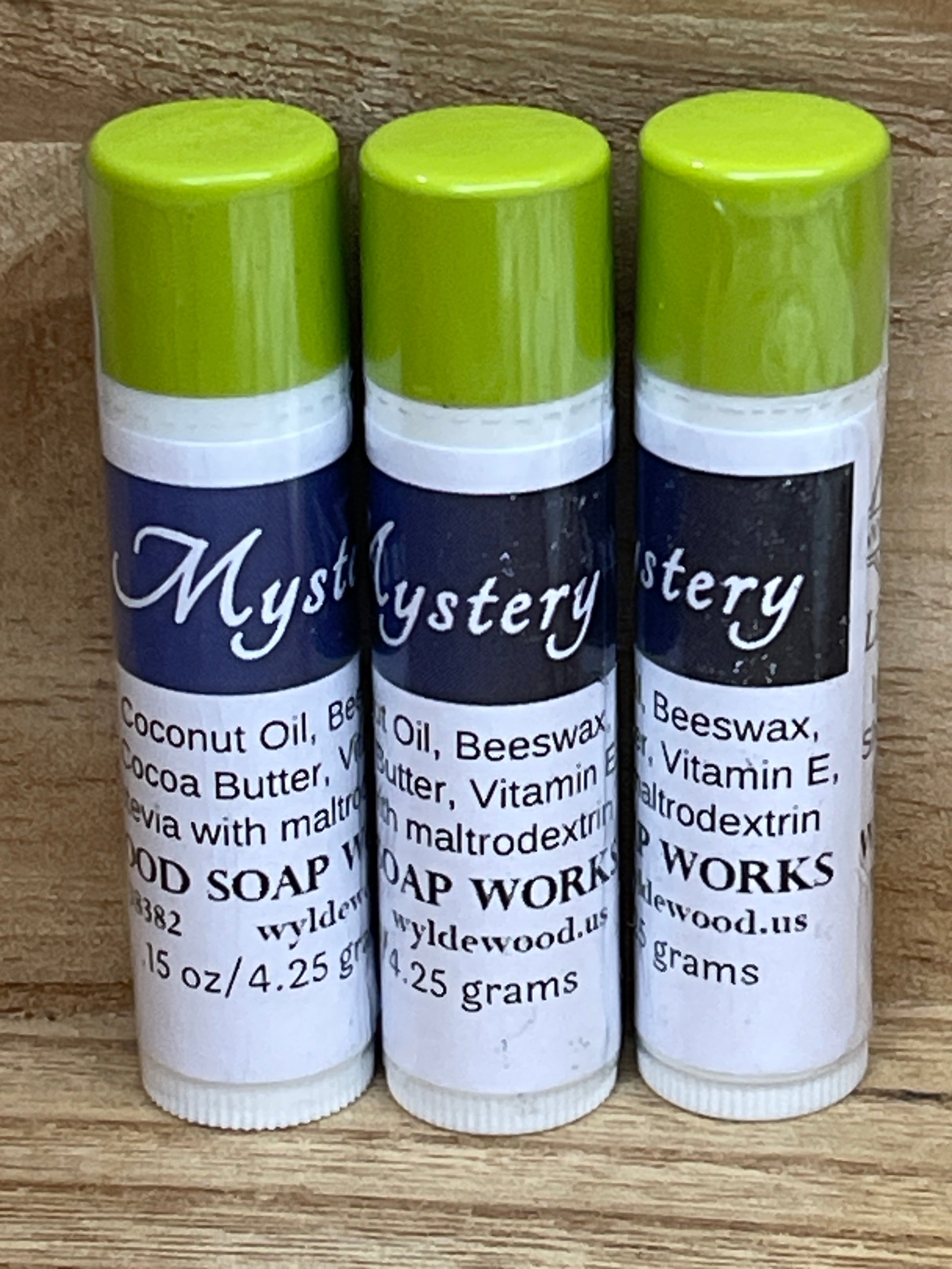 Three tubes of Mystery flavor lip balms made by Wyldewood soap works standing on a wood background.