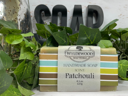 A bar of handmade goat milk soap with 'Patchouli' scent, placed on a white surface with green leaves and a 'SOAP' sign in the background.