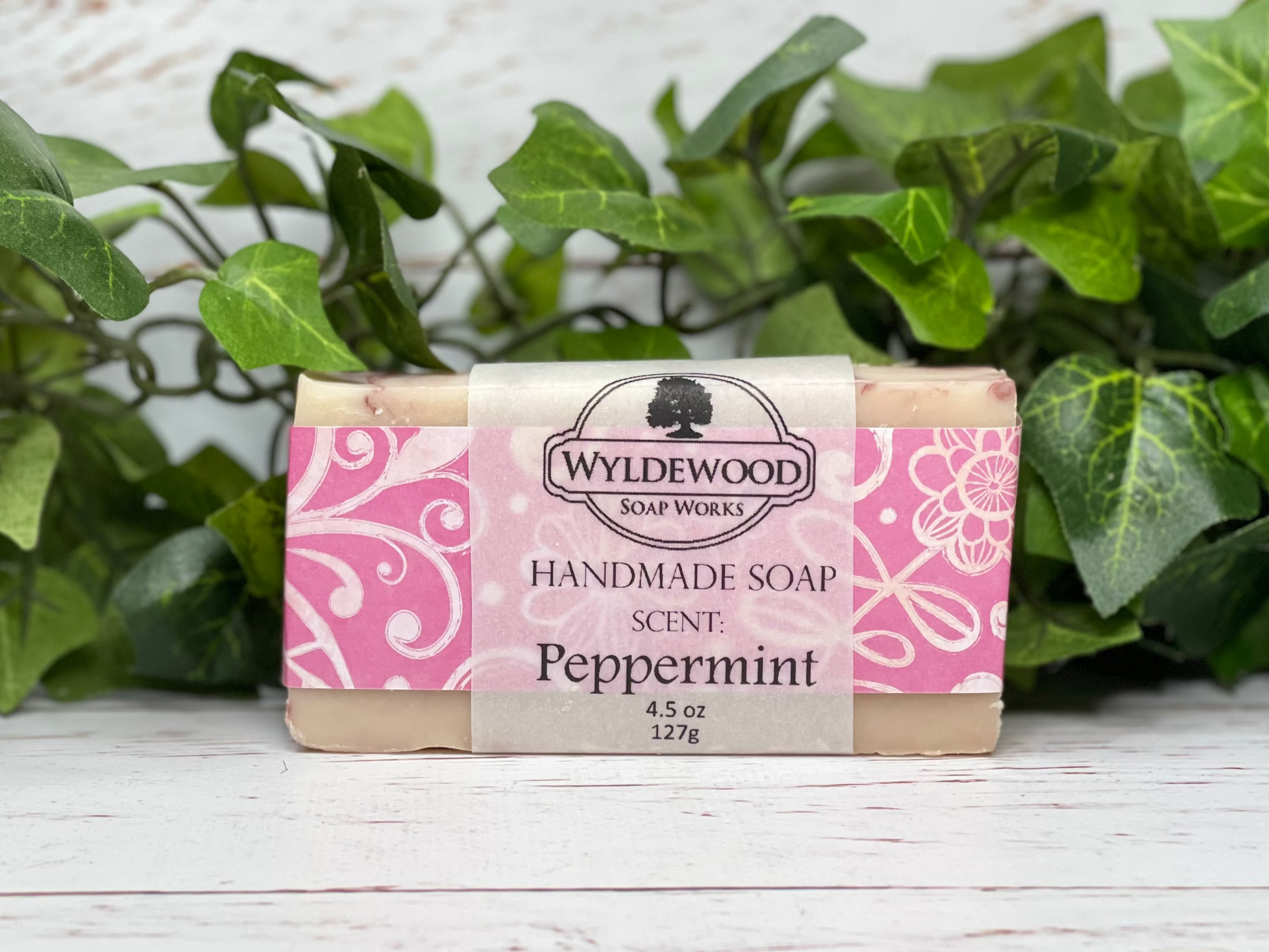 A bar of handmade soap with packaging that has 'WYLDEWOOD HANDMADE SOAP' and the scent listed as 'Peppermint'. The bar is placed on a white surface with leaves in the background.