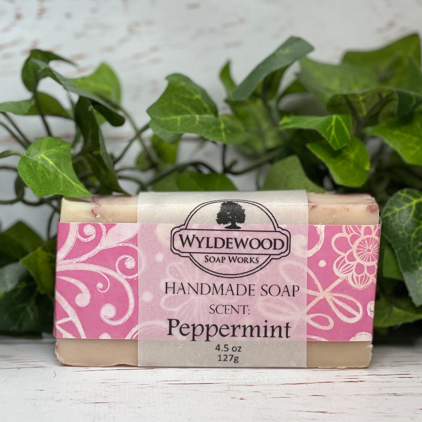 A bar of handmade soap with packaging that has 'WYLDEWOOD HANDMADE SOAP' and the scent listed as 'Peppermint'. The bar is placed on a white surface with leaves in the background.