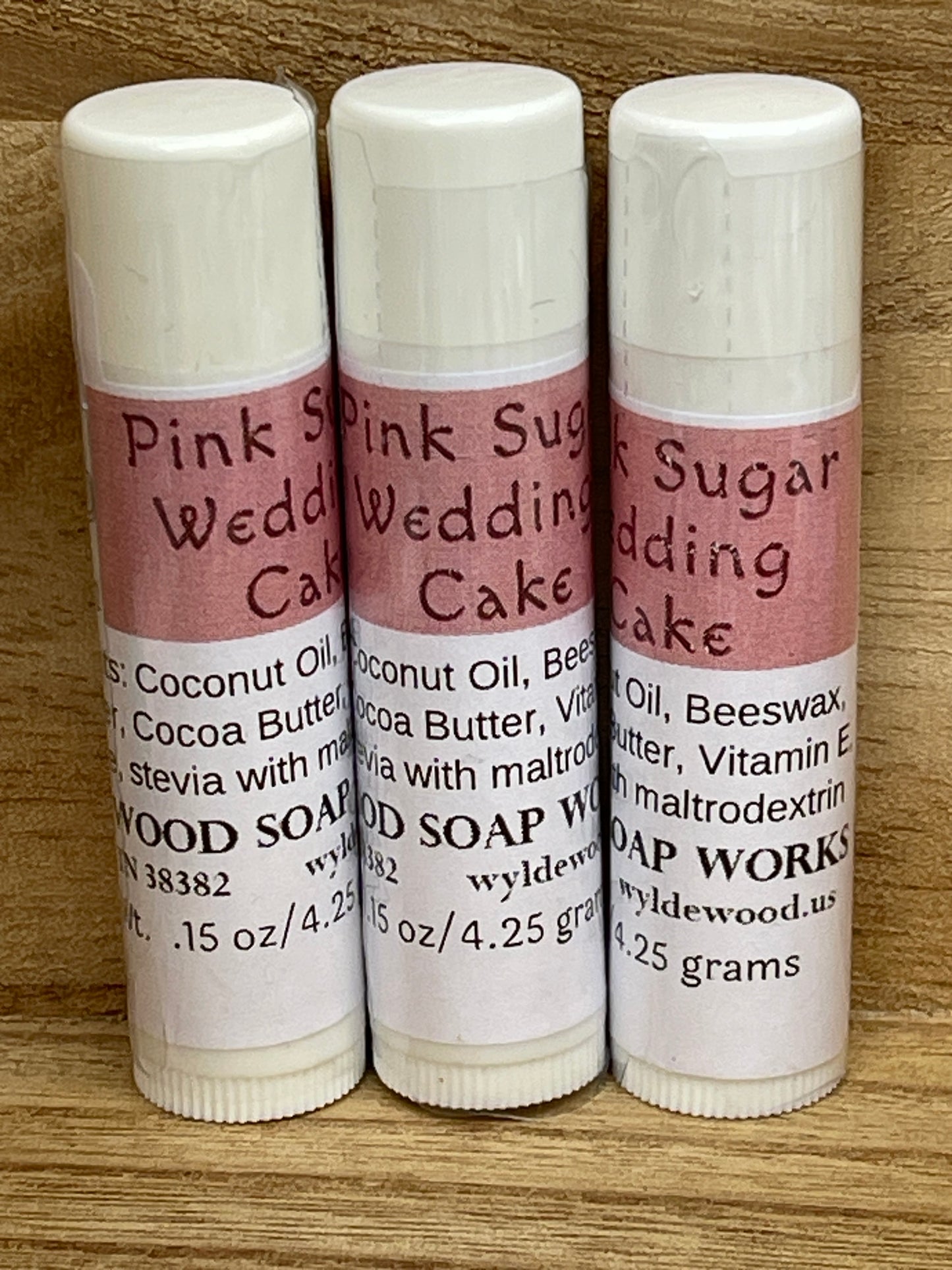 Three tubes of Pink Sugar Wedding Cake flavor lip balms made by Wyldewood soap works standing on a wood background.