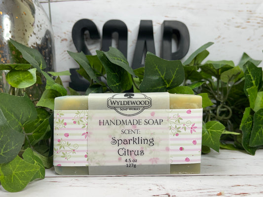 A bar of Wyldewood Soap Works Scented Handmade Soap in scent Sparking Citrus on a white background with greenery and a sign that says "Soap" behind the bar of soap.