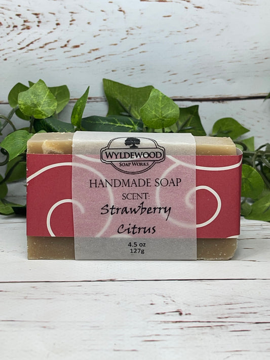 A bar of handmade soap with packaging that has 'WYLDEWOOD HANDMADE SOAP' and the scent listed as 'Strawberry Citrus'. The bar is placed on a white surface with leaves in the background.