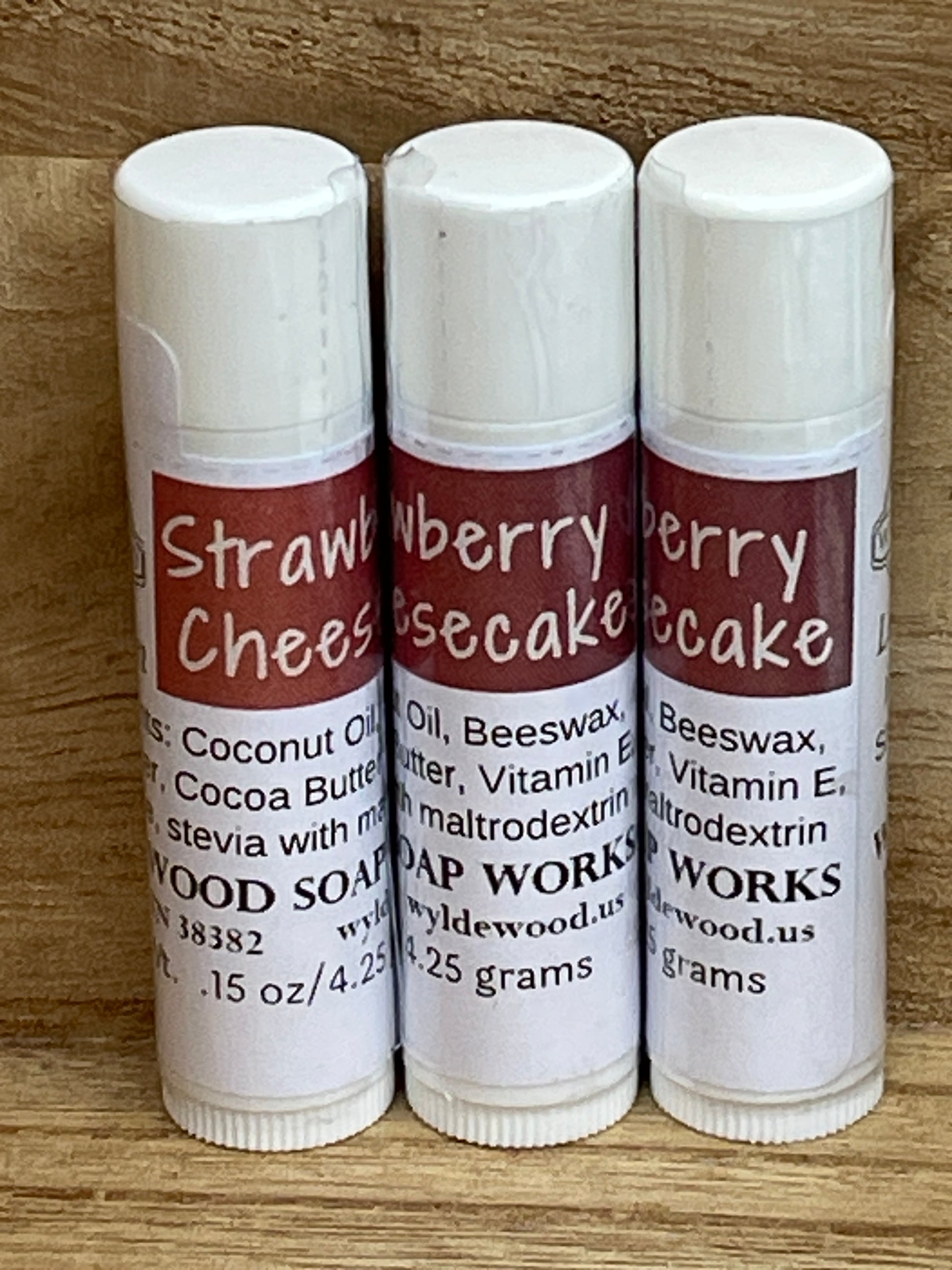 Three tubes of Strawberry Cheesecake flavor lip balms made by Wyldewood soap works standing on a wood background.