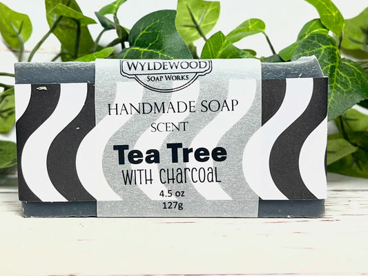 Handmade soap bar with 'Tea Tree with Charcoal' scent packaging, Wyldewood Soap Works brand, on a white background with green leaves.