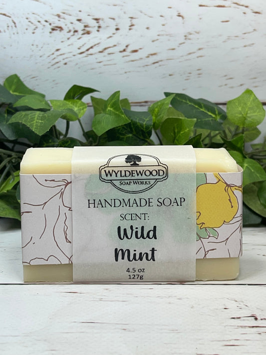 A light cream colored bar of scented goat milk soap named Wild Mint wrapped with yellow and green pattered scrapbook paper and a vellum label on a white back ground with green leaves behind in the background